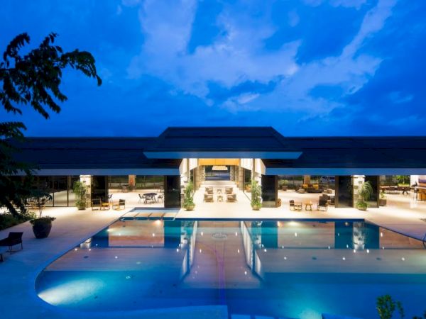A luxurious, modern building with a large, illuminated swimming pool in the foreground, set against a dramatic evening sky.