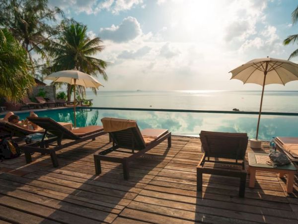 A serene poolside scene with lounge chairs, sun umbrellas, and a view of the ocean under a partly cloudy sky.