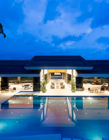 A modern building with large windows is illuminated at night, overlooking a spacious, brightly lit swimming pool.