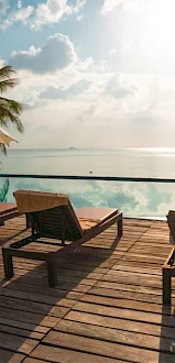A wooden deck with loungers and umbrellas overlooks an infinity pool and ocean under a partly cloudy sky.