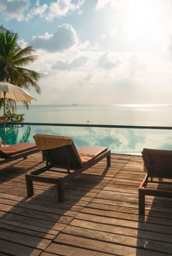 Sunlit poolside with lounge chairs and umbrellas overlooking a calm ocean, surrounded by palm trees under a partly cloudy sky.