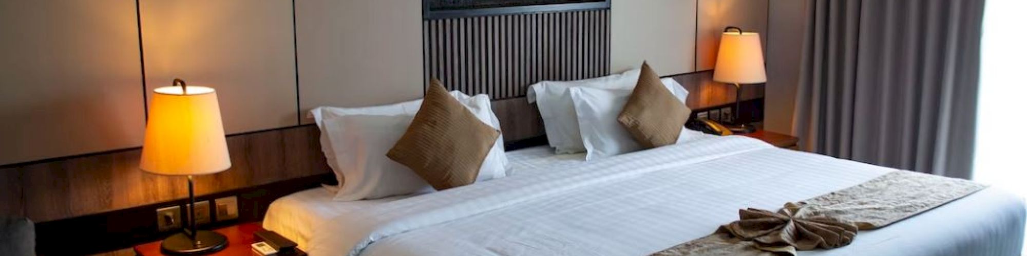 A modern hotel room with a large bed, side tables, lamps, and artwork above the bed. Curtains cover a window on the right side.