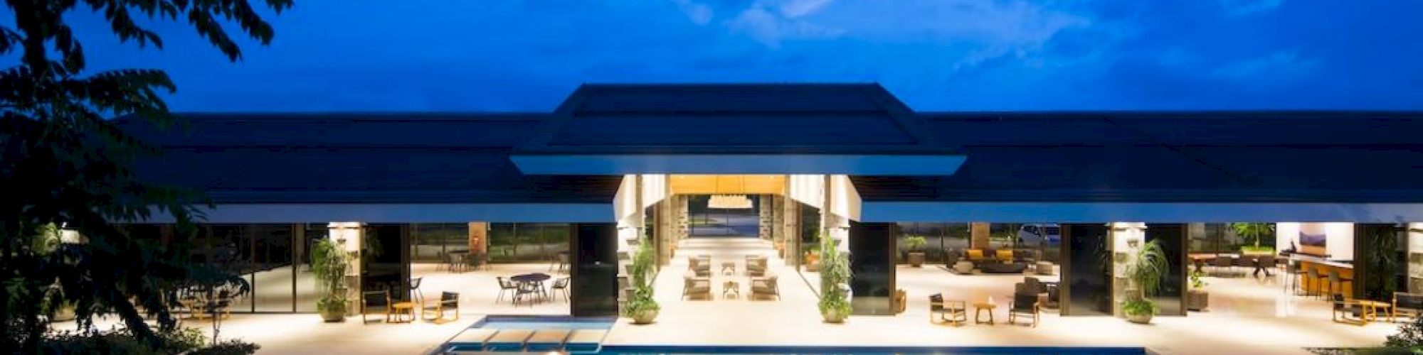 A modern building with open spaces and glowing lights overlooks a large swimming pool under a deep blue evening sky.