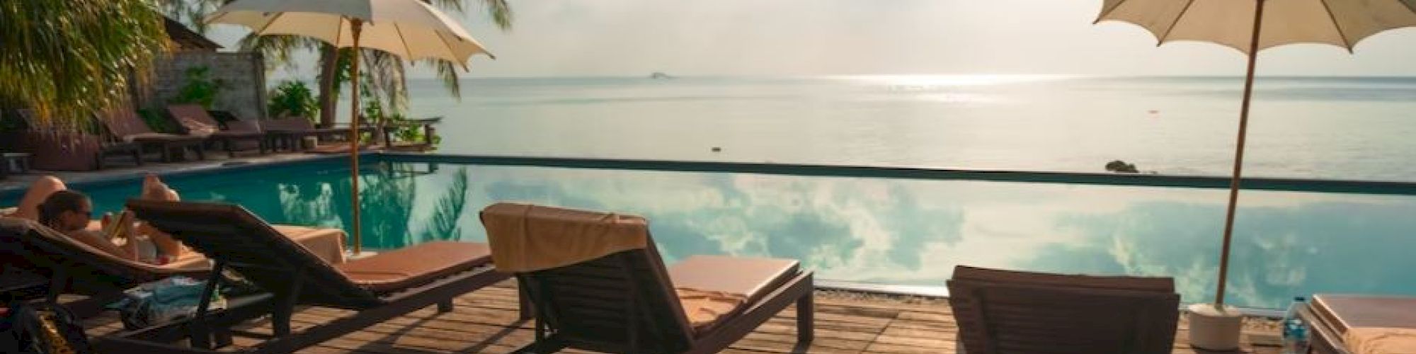 A serene poolside view with lounge chairs, umbrellas, and palm trees overlooking a calm ocean under a partly cloudy sky.