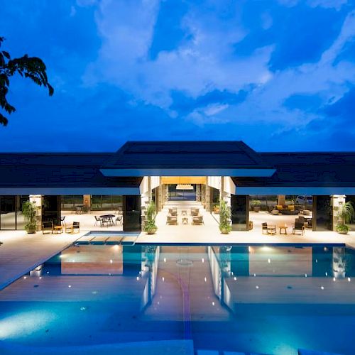 A luxurious modern building with a large illuminated pool at dusk, surrounded by lounge chairs and greenery under a blue sky.