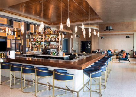 The image shows a modern bar with a wooden countertop, stylish stools, hanging lights, and a wide array of liquor bottles on shelves.