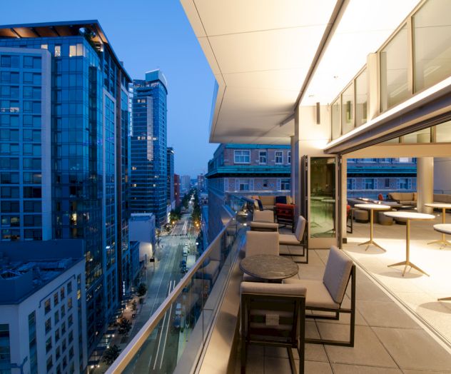 A modern cityscape with tall buildings at dusk and a sleek, furnished balcony overlooking the street below.