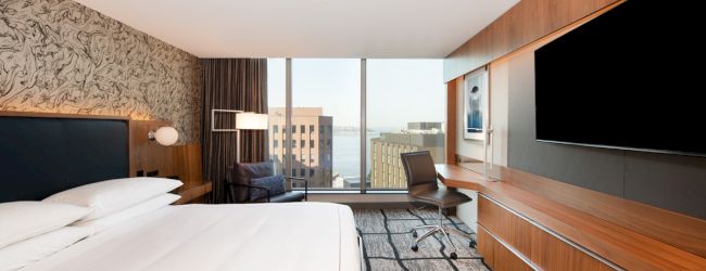 A modern hotel room with a large bed, desk, chair, and a wall-mounted TV. The window provides a view of buildings and a glimpse of water.