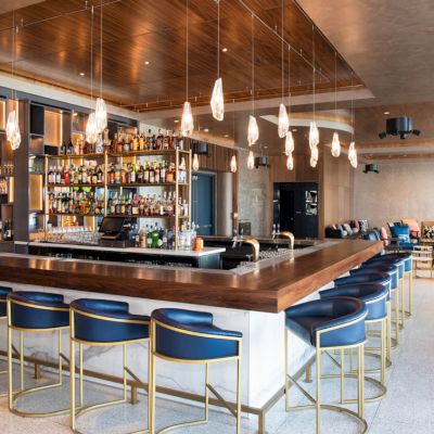 This image shows a stylish bar with a wooden countertop, blue cushioned stools, and hanging lights, surrounded by shelves of bottles.