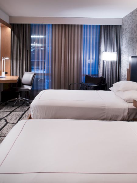 A modern hotel room with two beds, a flat-screen TV, a desk, and stylish lighting and decor.