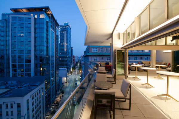 A modern rooftop lounge overlooks a cityscape with skyscrapers at dusk, featuring seating, tables, and soft lighting on the terrace.