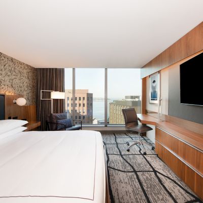 The image shows a modern hotel room with a large bed, a work desk, a wall-mounted TV, and a window with a cityscape view.