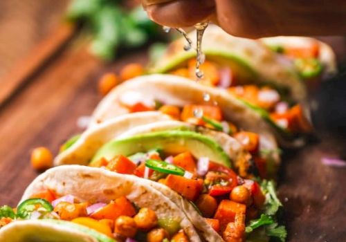 A hand squeezes lime juice over a row of colorful veggie tacos with avocado, chickpeas, and toppings, all on a wooden board.