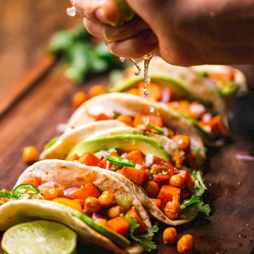 A hand squeezes lime juice over a row of colorful veggie tacos with avocado, chickpeas, and toppings, all on a wooden board.