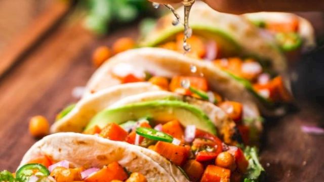 A hand squeezes lime juice over a row of colorful veggie tacos with avocado, chickpeas, and toppings, all on a wooden board.