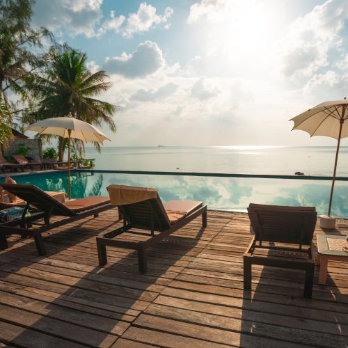 A scenic view of a tropical beachside pool area, featuring lounge chairs, umbrellas, and palm trees with a calm ocean in the background.
