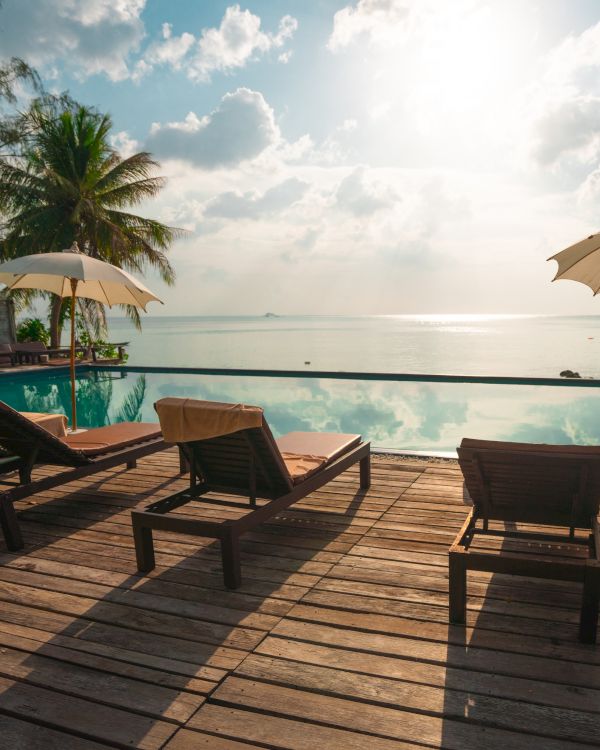 A scenic view of a tropical beachside pool area, featuring lounge chairs, umbrellas, and palm trees with a calm ocean in the background.