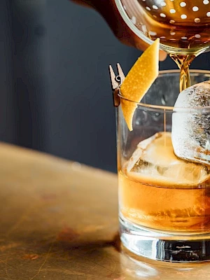 A cocktail is being poured into a glass with ice and a twist of lemon peel on the side, placed on a bar counter.