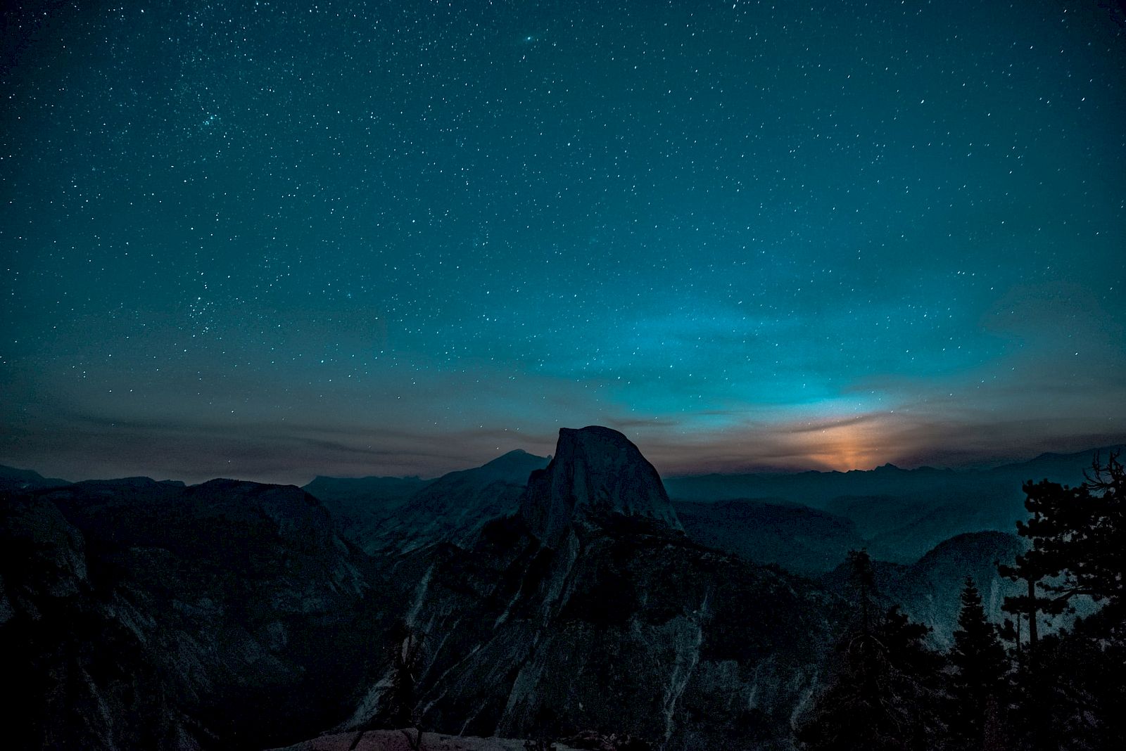 A starry night sky over a mountainous landscape with trees, featuring a prominent peak and a glowing horizon.