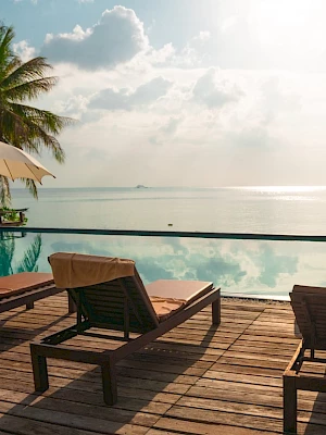 A serene poolside scene with lounge chairs, sun umbrellas, and a view of the ocean under a partly cloudy sky. Palm trees are in the background.