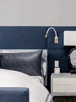A modern bedroom setup featuring a navy headboard, white bed, side table with a lamp, and wall-mounted reading light.