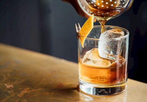 A cocktail being poured into a glass with a large ice cube and a twist of citrus peel garnish on a wooden surface.