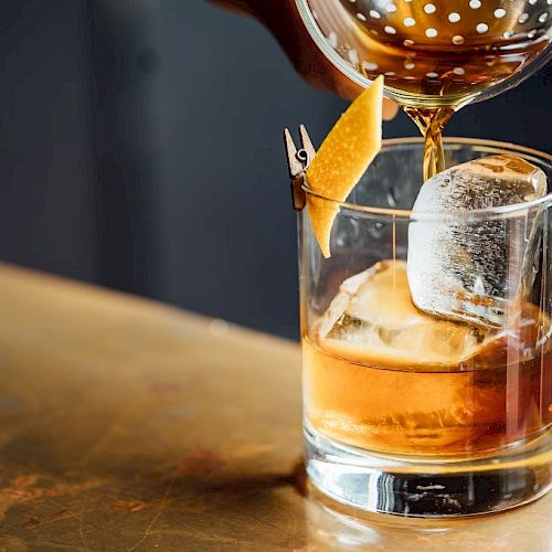 A cocktail being poured into a glass with a large ice cube and a twist of citrus peel garnish on a wooden surface.