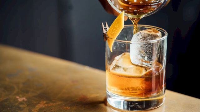 A cocktail being poured into a glass with a large ice cube and a twist of citrus peel garnish on a wooden surface.