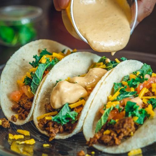 Three tacos filled with ground meat, lettuce, cheese, and tomatoes, being topped with sauce on a plate.