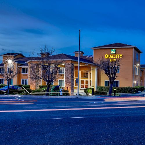 The image shows a Quality Inn hotel building with a well-lit exterior at dusk, cars parked in front, and light trails on the street.
