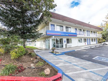A two-story motel with a "Welcome" sign, parking spaces, and surrounding greenery under a partly cloudy sky.