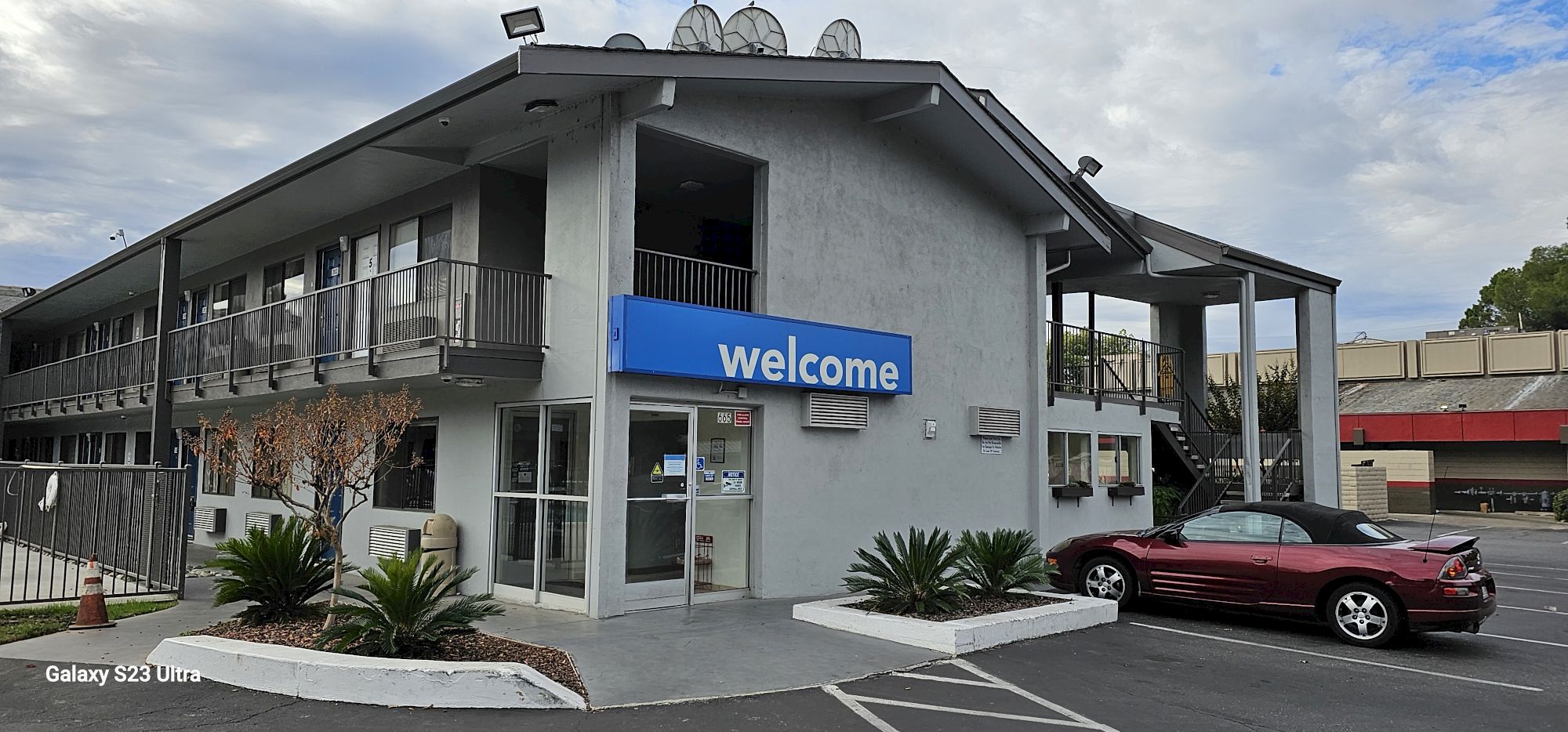 A motel with a "welcome" sign, a red convertible car parked in front, and a two-story building with exterior corridors.