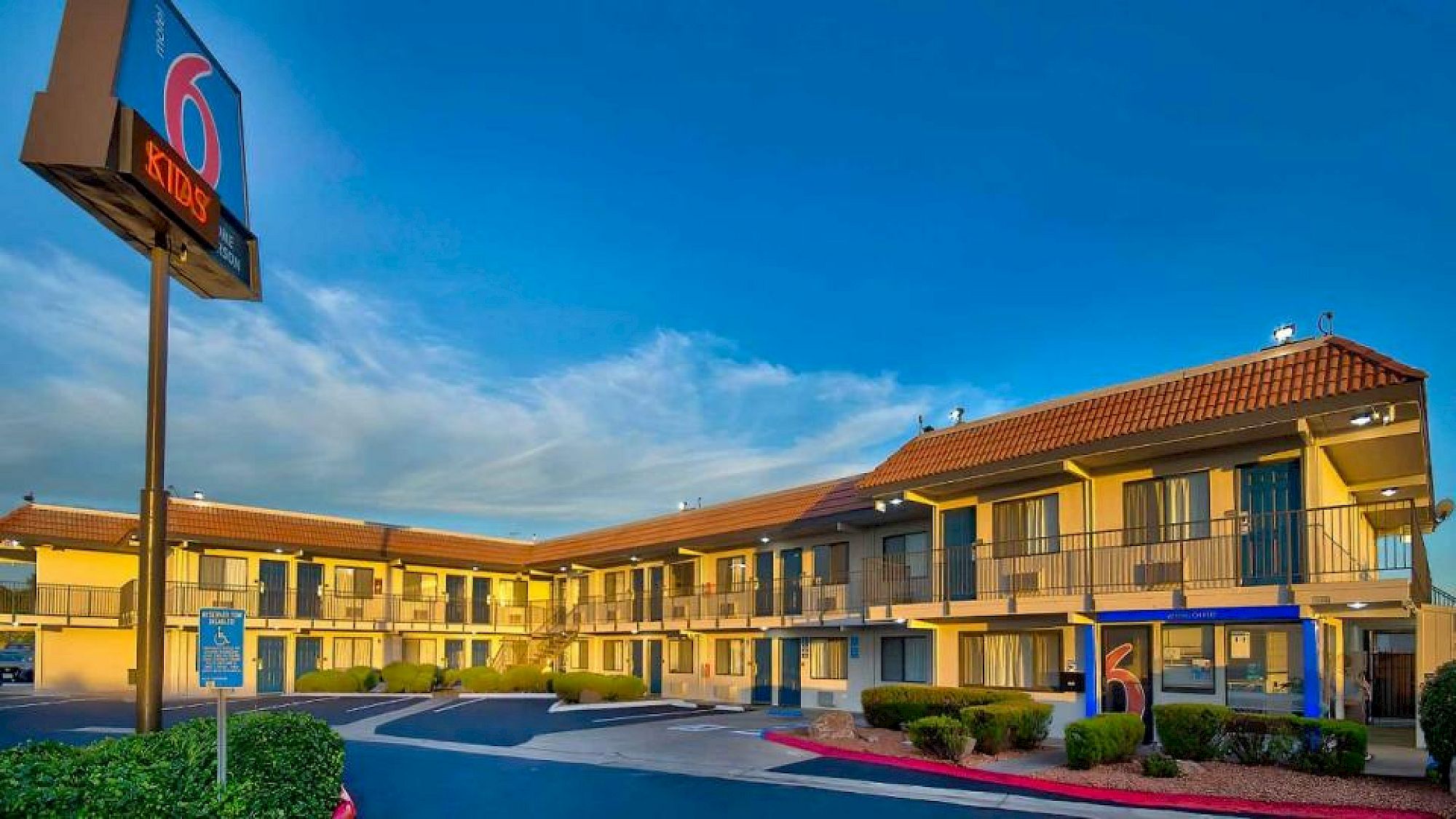 A two-story motel with a sign displaying the number "6" is shown against a clear blue sky, surrounded by neatly trimmed bushes and a parking area.