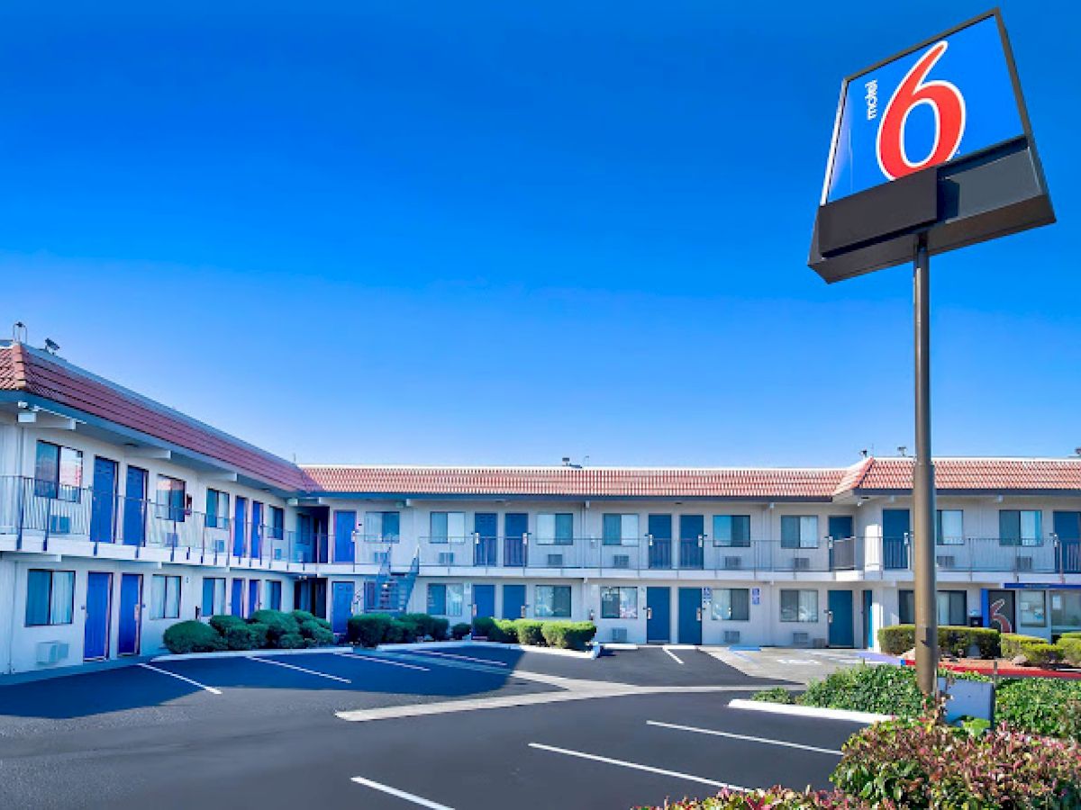 The image shows a two-story motel with a "6" sign, featuring an outdoor parking area, blue doors, and a well-maintained exterior.