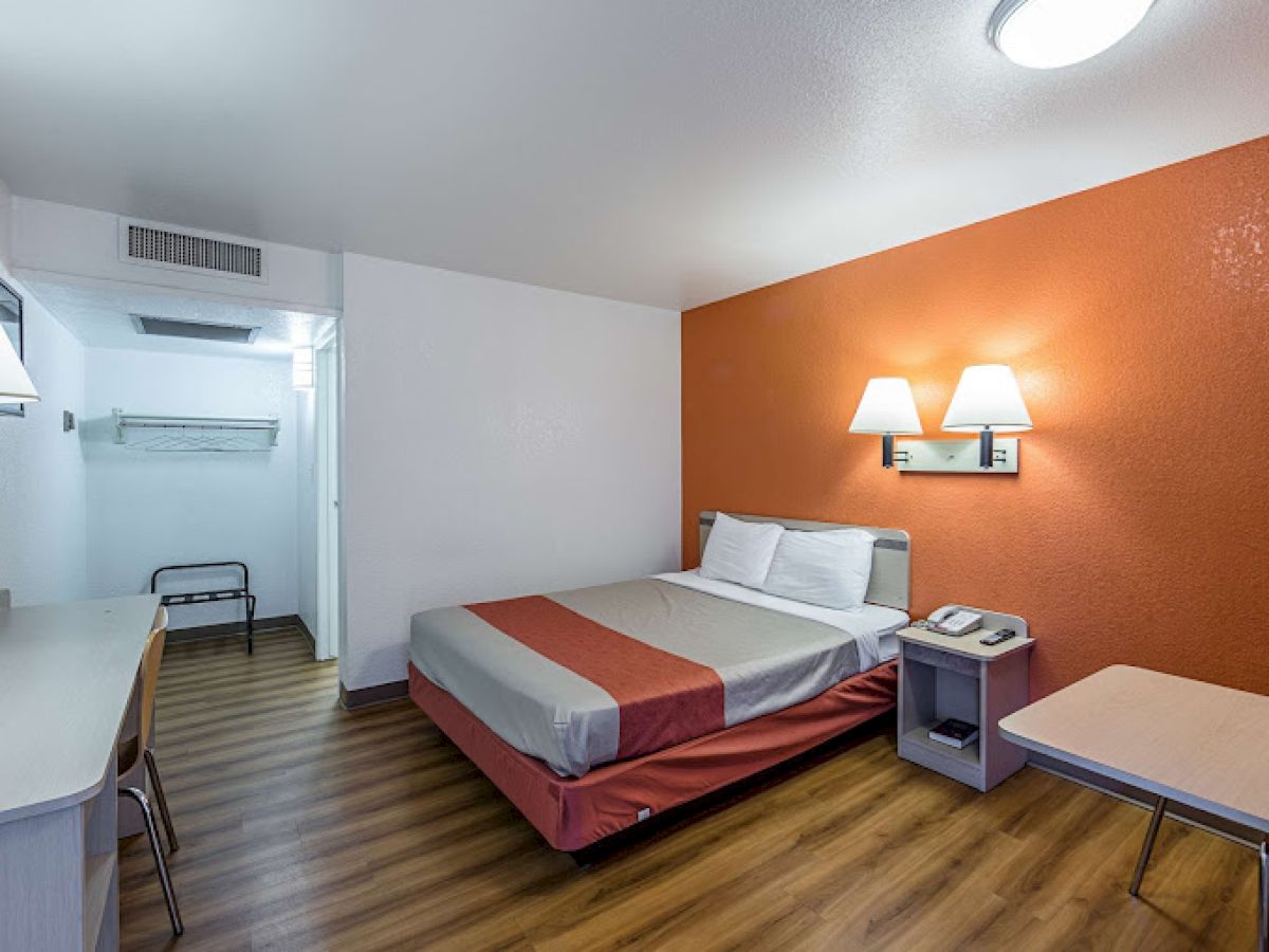 A simple hotel room with a bed, desk, phone, lamps, and wooden floor. Walls are white and orange. Modern and minimal décor.