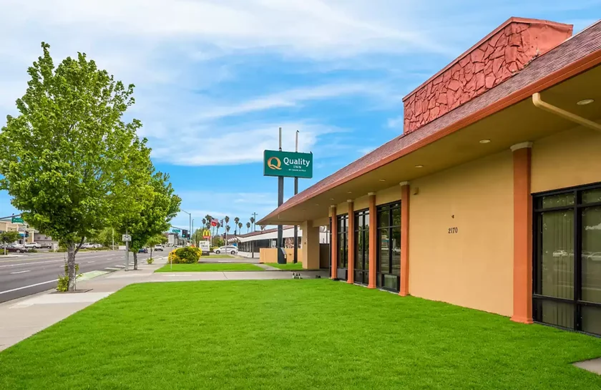 A roadside building with a "Quality Inn" sign, green grass, and blue sky. The street is lined with trees and flags.