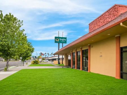 A street view with a grassy lawn, trees, and a building with a sloped roof; a "Quality Inn" sign is visible in the background.