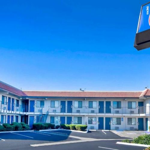 A motel with a "6" sign, featuring a two-story building, parking area, and blue doors. The sky is clear and bright blue.