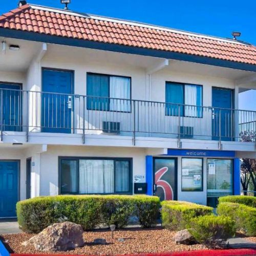 A two-story motel with blue doors, a red-tiled roof, and a "6" logo on the exterior, surrounded by shrubs and a parking space.