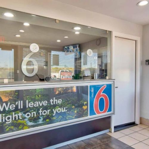A reception area with a Motel 6 logo, a counter, and the phrase "We'll leave the light on for you." The space is well-lit and tidy.