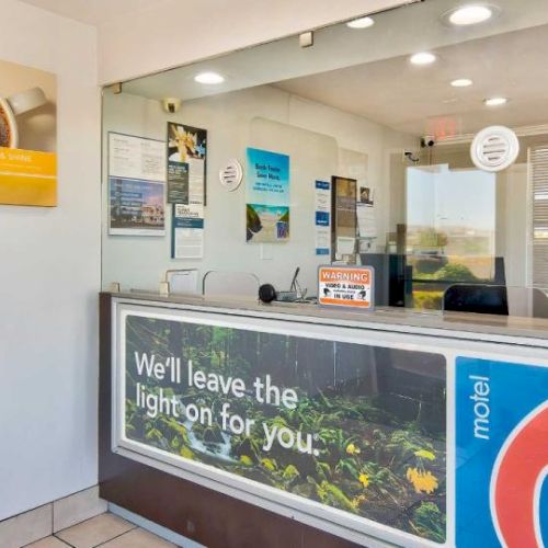 The image shows a Motel 6 reception area with signs, brochures, and a counter with a protective screen. The slogan "We'll leave the light on for you."