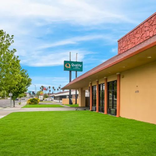 The image shows a commercial building with a lawn, a tree, and a sign for "Quality" against a clear blue sky on a city street corner.