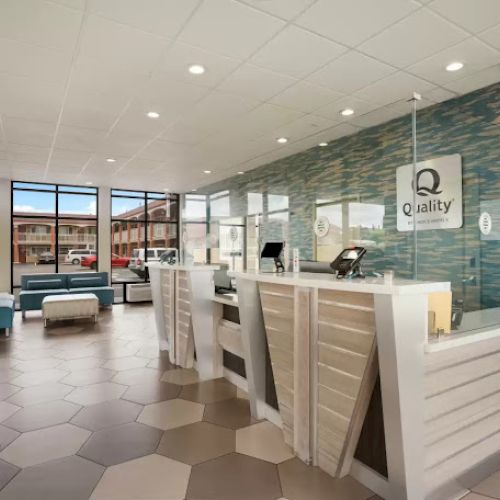 A modern hotel lobby with hexagonal floor tiles, a front desk, and a "Quality" sign on the wall. Bright and welcoming atmosphere.