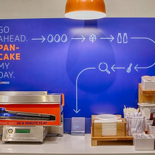 This image shows a pancake-making station with a conveyor oven, a sign saying "PANCAKE MY DAY," and various toppings and utensils.