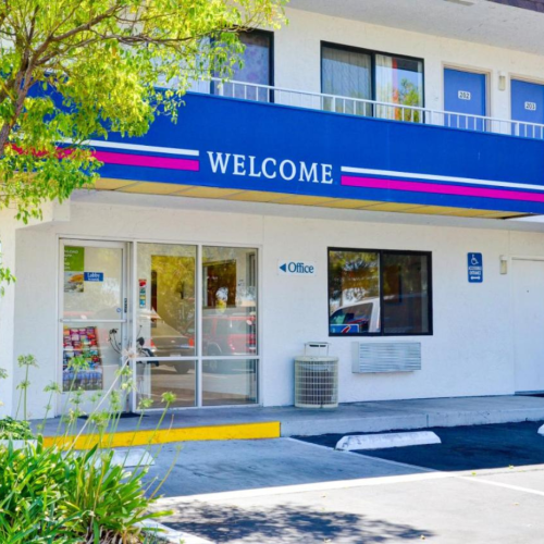 The image shows the entrance to a motel with a sign saying "WELCOME," featuring a glass door labeled "Office" and a second floor with room doors.