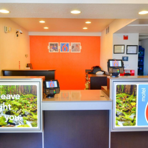 The image shows the reception area of a Motel 6 with a bright orange wall and signage saying "We'll leave the light on for you."