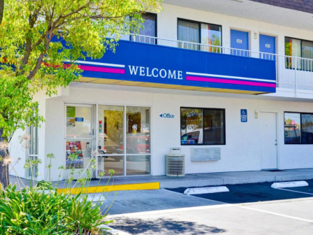 The image shows the entrance of a motel with a "WELCOME" sign, doors labeled with room numbers, and greenery in the foreground.