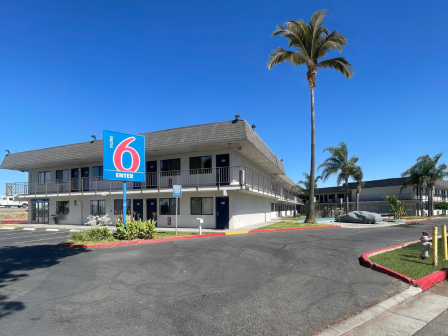 A motel with a "6" sign, palm trees, clear blue sky, and a two-story building with balconies in a parking lot.