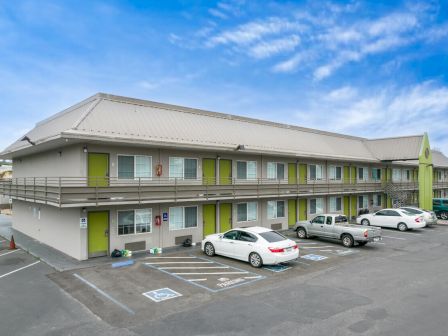 The image shows a two-story motel with a parking lot in front, featuring several parked cars and a modern architectural design.
