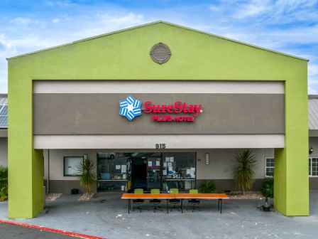 The image shows the entrance of a SureStay Plus Hotel, featuring a green and gray facade with a sign above the door, and outdoor seating.